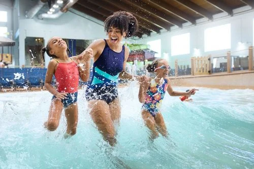 A smiling woman and two young girls in swimsuits are playing in the water at an indoor waterpark, creating a joyful and playful scene.