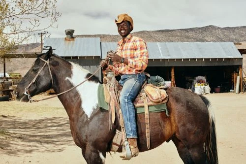 Man riding a brown and white horse in an outdoor rural setting with a building and distant mountains visible under a bright sky.