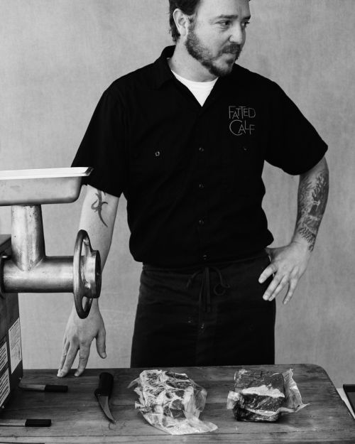 Man with beard stands at a table with meat grinder, raw meat, knife and wooden cutting board in a black and white studio shot.