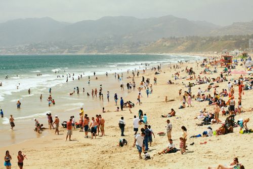 Wide shot of a crowded beach with many people enjoying the sand and sea under a bright, overcast sky, backed by distant mountains.