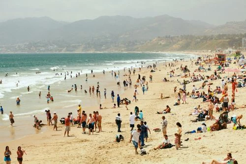 Wide shot of a crowded beach with many people enjoying the sand and sea under a bright, overcast sky, backed by distant mountains.
