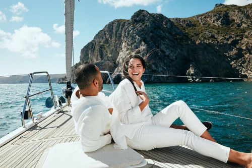 Couple in white clothing relax on a sailboat under a partly cloudy sky with a rock island backdrop, enjoying a sunny day at sea.