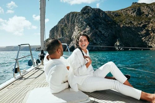 Couple in white clothing relax on a sailboat under a partly cloudy sky with a rock island backdrop, enjoying a sunny day at sea.