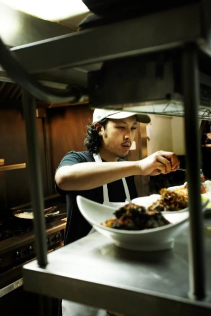 Adult male chef in a baseball cap plates food in a restaurant kitchen with stainless steel surfaces, shelves, and industrial cooking equipment.