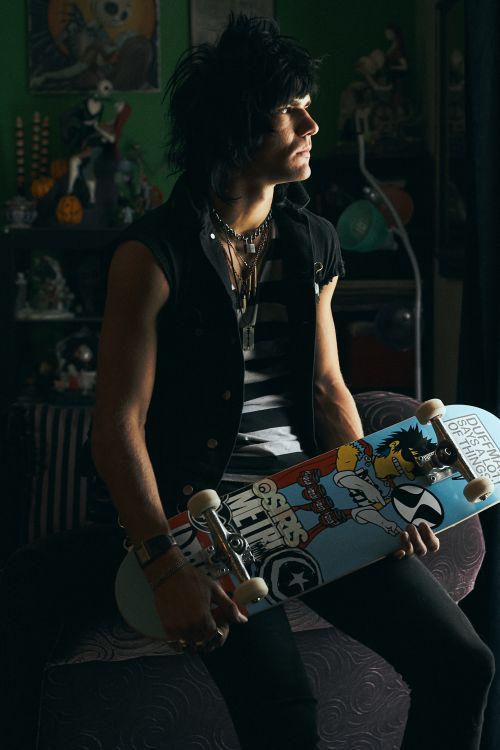 Young adult with dark hair, sitting indoors, holding a skateboard with graphic art, wearing a vest, striped shirt, and dark pants.