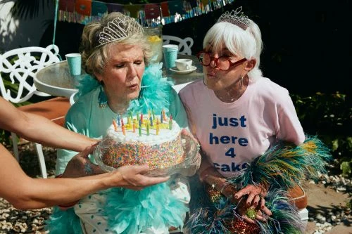 Two elderly women in crowns celebrate a birthday outdoors with a sprinkle-covered cake and turquoise feather boas.