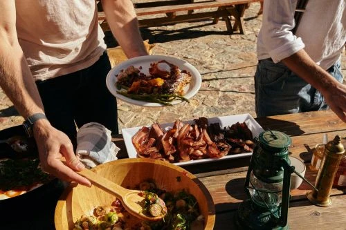 Close-up on a rustic wood table with food, plates, wooden salad bowl, lantern, and two people serving food during an outdoor meal.