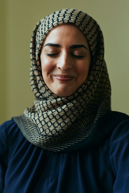 Portrait of a woman wearing a patterned hijab, eyes closed, with a slight smile, against a plain background in soft, diffused lighting.