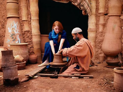Woman and man work at a pottery wheel indoors surrounded by earthenware vessels, showcasing the creation of ceramics in a rustic setting.
