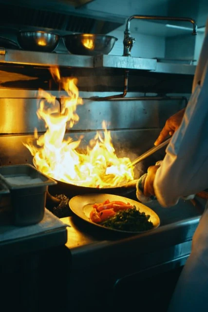 A chef in a restaurant kitchen is cooking food in a frying pan with flames, with a plate of cooked vegetables nearby on a steel counter.