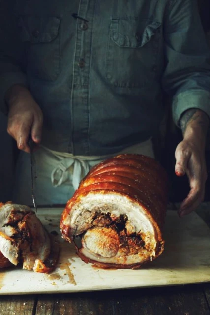 Rolled pork roast being sliced by an adult in a blue shirt, on a wooden board, in dim, natural lighting for a food advertisement.
