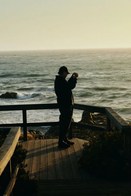 A man in silhouette stands on a wooden platform by the ocean, taking a picture with his phone on a clear day, overlooking the water.