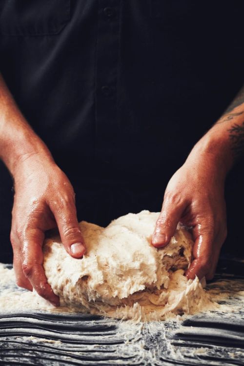 Close-up of a person kneading dough on a dark surface, wearing a black shirt, capturing a moment of food preparation.