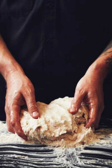 Close-up of a person kneading dough on a dark surface, wearing a black shirt, capturing a moment of food preparation.