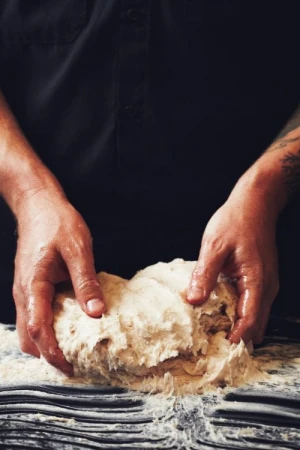 Close-up of a person kneading dough on a dark surface, wearing a black shirt, capturing a moment of food preparation.