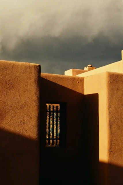 Exterior architecture with textured adobe walls, dark window and shadows cast on the surface under a cloudy sky on the horizon.