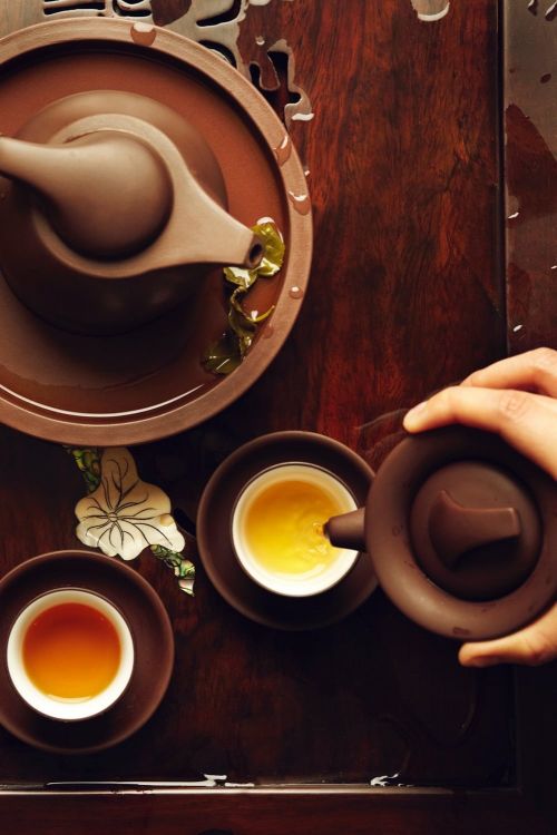 Overhead shot of a tea ceremony, featuring a hand pouring tea from a brown teapot into a small white cup with brown saucers.