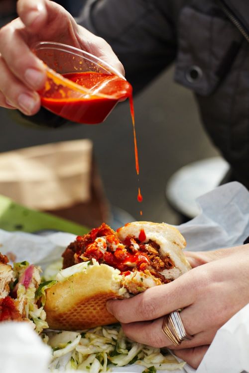 Close-up of a person pouring red sauce onto a chicken sandwich with white coleslaw, showcasing food preparation and handheld cuisine.