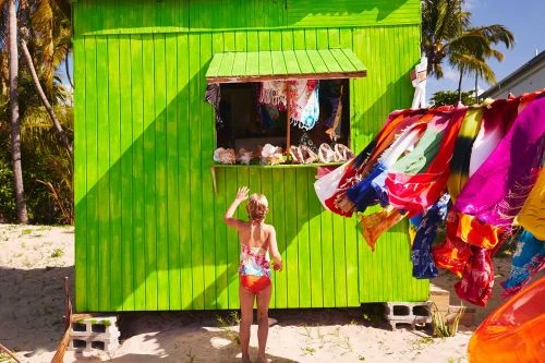A young girl in a swimsuit stands near a brightly colored green shop with colorful textiles hanging outside on a sunny, tropical beach.