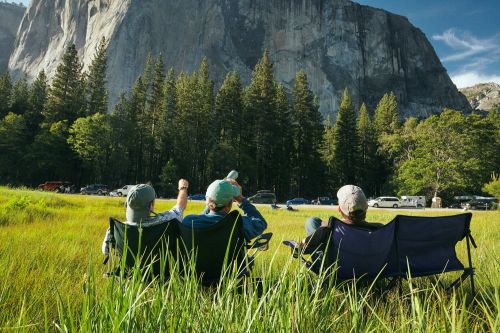 Three people sit in chairs in a grassy meadow with a mountain in the background on a sunny day, enjoying the scenic view.