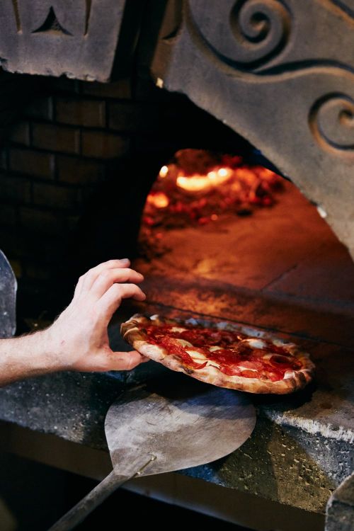 Close-up of a pizza being placed into a brick oven by a cook using a metal peel, with fire visible inside the oven.