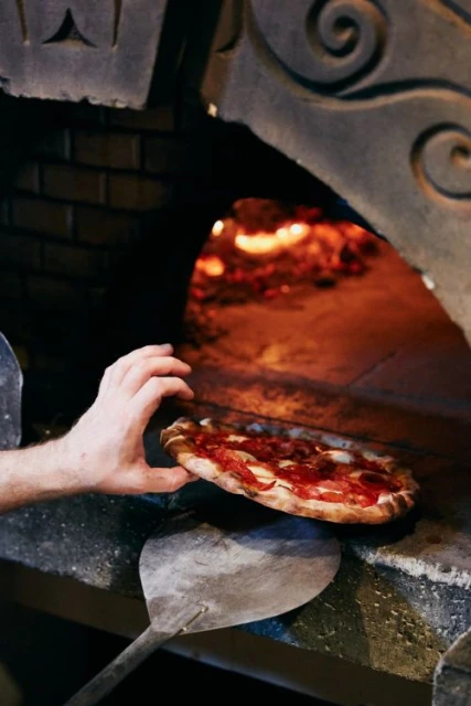 Close-up of a pizza being placed into a brick oven by a cook using a metal peel, with fire visible inside the oven.