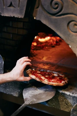 Close-up of a pizza being placed into a brick oven by a cook using a metal peel, with fire visible inside the oven.