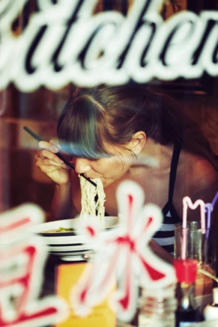 An adult woman eats noodles with chopsticks while sitting indoors; food and restaurant window signage are in the foreground.