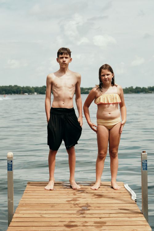 Two children stand on a wooden dock, facing forward, in bathing suits on a sunny day. Water and trees are visible in the background.