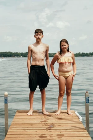 Two children stand on a wooden dock, facing forward, in bathing suits on a sunny day. Water and trees are visible in the background.