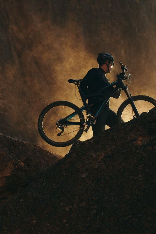 A man carries a mountain bike up a rocky hill with a dramatic, hazy backdrop in a low-angle, medium shot, capturing an outdoor, sporty scene.