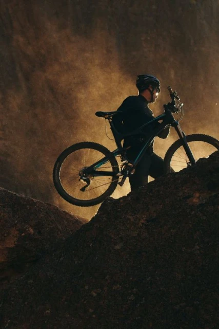 A man carries a mountain bike up a rocky hill with a dramatic, hazy backdrop in a low-angle, medium shot, capturing an outdoor, sporty scene.