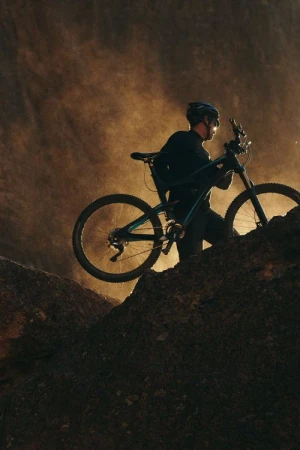 A man carries a mountain bike up a rocky hill with a dramatic, hazy backdrop in a low-angle, medium shot, capturing an outdoor, sporty scene.