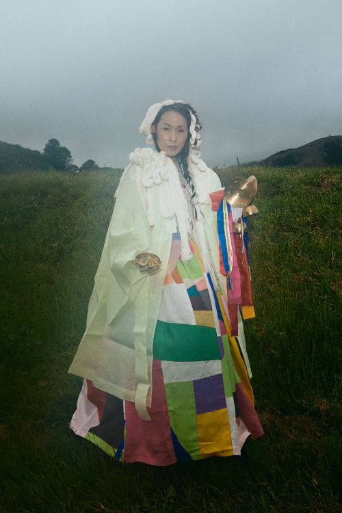A woman in a colorful patchwork gown stands in a green field with a hazy sky, holding a golden object. The overall scene is artistic.
