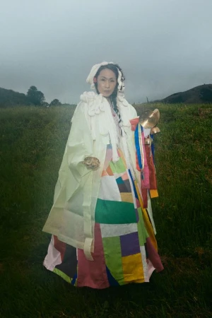 A woman in a colorful patchwork gown stands in a green field with a hazy sky, holding a golden object. The overall scene is artistic.
