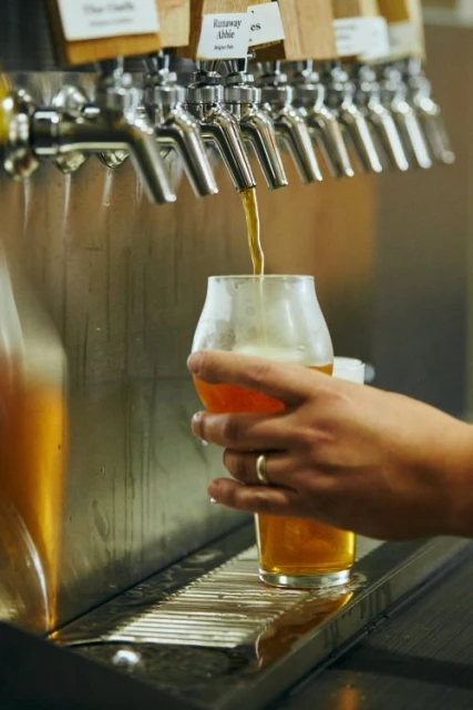 Close-up of a person pouring amber-colored beer from a stainless steel tap into a clear tulip glass at a bar.