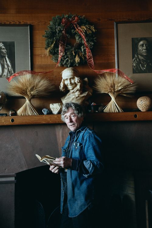 Adult man with gray hair stands indoors in front of a mantle and reads a book in a warmly lit room. Wreath, busts, and art decorate the wall.
