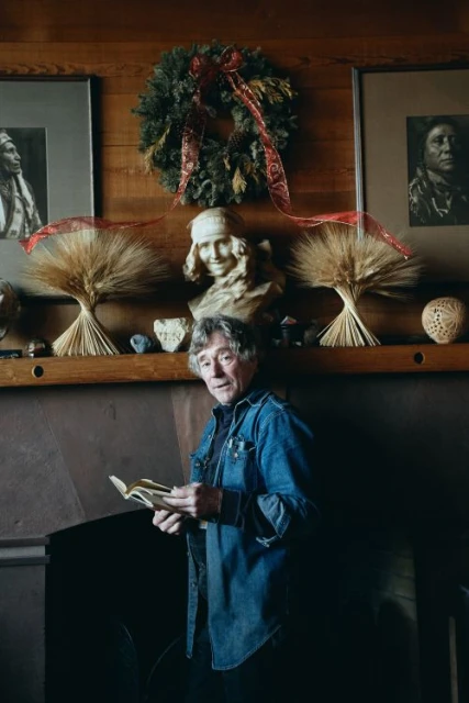 Adult man with gray hair stands indoors in front of a mantle and reads a book in a warmly lit room. Wreath, busts, and art decorate the wall.