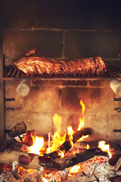 Grilled ribs over an open fire. Close-up showcases the cooking meat atop a black grate, with bright flames and glowing embers below.