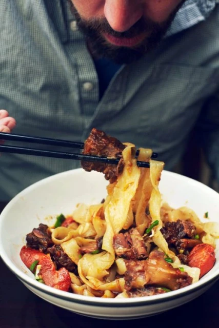 Close-up of a bearded man eating noodles with beef and vegetables from a white bowl using chopsticks, showcasing a savory meal.