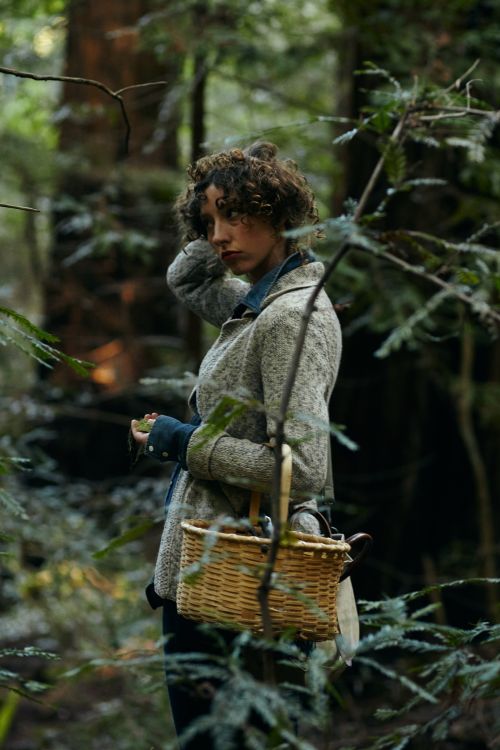 Woman with curly brown hair wearing a tweed jacket holding a woven basket in a dense redwood forest.
