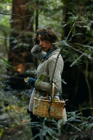 Woman with curly brown hair wearing a tweed jacket holding a woven basket in a dense redwood forest.