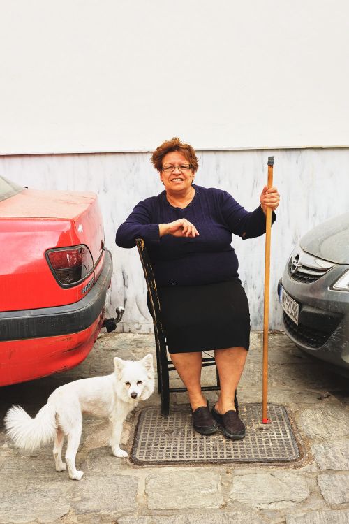 Elderly woman sitting outdoors with her white dog, flanked by parked cars on a sunny day. She holds a cane and smiles.