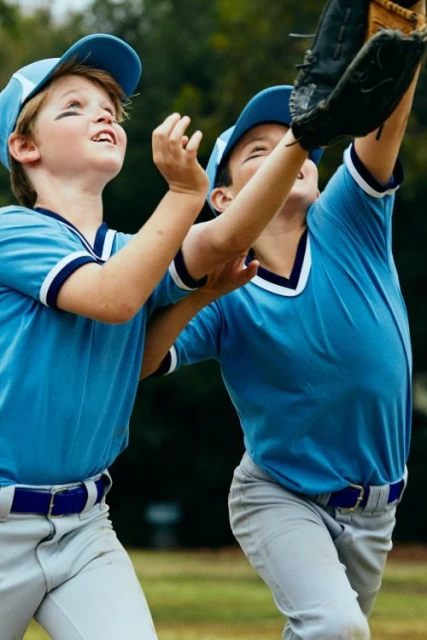 Two young boys in baseball uniforms reaching for a ball on a grassy field, wearing caps and gloves, with trees in the background.