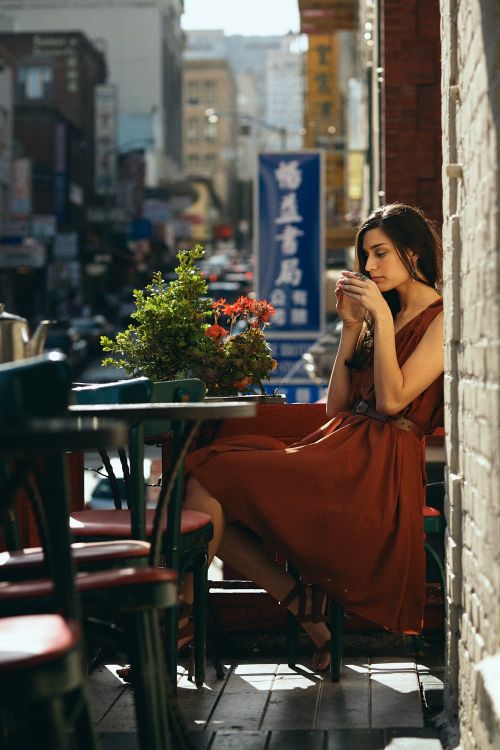 A young woman in a rust-colored dress sips from a cup at an outdoor cafe in Chinatown, with a street scene in the background.