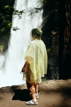 Person in a yellow raincoat stands viewing a waterfall in a forest; trees and greenery frame the background of the natural scene.