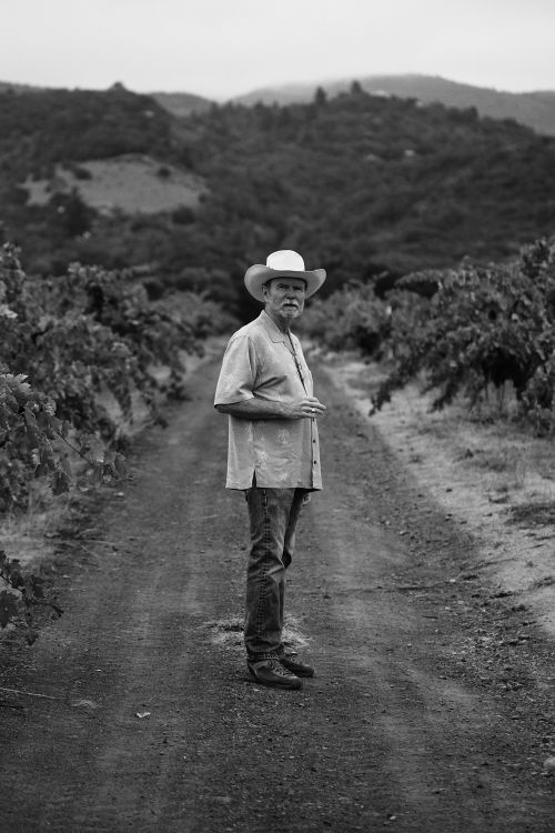 Man in cowboy hat stands centered in vineyard with mountain backdrop in black and white, wearing jeans and button-down shirt.
