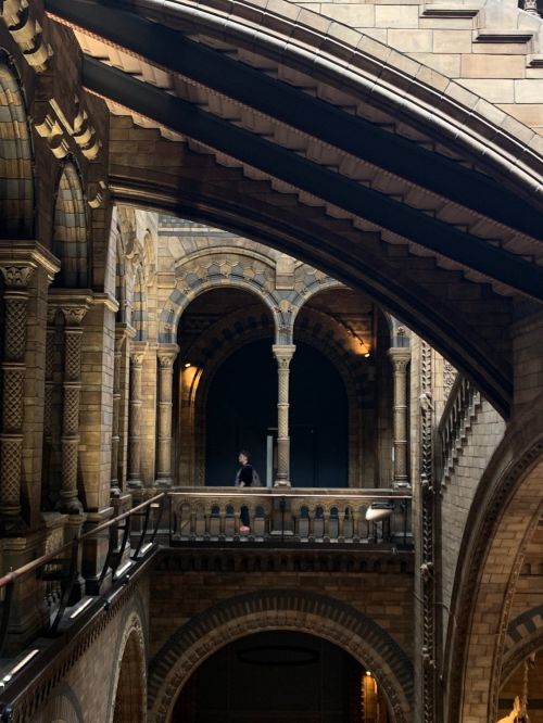Architectural interior of a grand building featuring arched walkways, stone walls, and two people standing on a balcony.