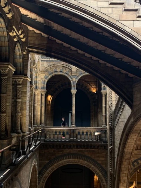Architectural interior of a grand building featuring arched walkways, stone walls, and two people standing on a balcony.