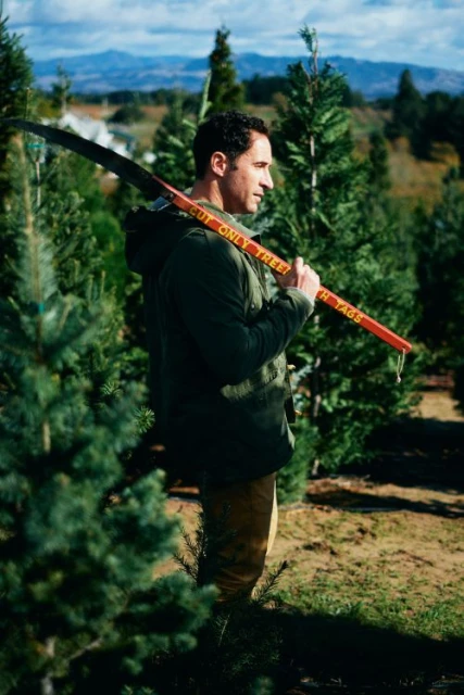 An adult male carries a tree trimming tool with an orange handle in a Christmas tree farm on a sunny day with mountains in the background.